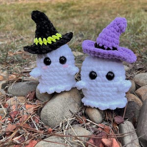 May include: Two white crocheted ghost figures with black plastic eyes and pink stitched cheeks. One ghost wears a black pointed hat with a neon yellow band, and the other wears a light purple pointed hat with a black band, both made of yarn.