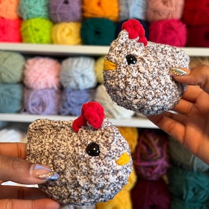 May include: Two crocheted gray and white chicks with red and yellow accents. The chicks are sitting on a shelf with colorful yarn balls in the background.