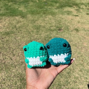 May include: Two crocheted dinosaur plushies. One is bright green, the other is teal. Both have white bellies and black button eyes. The plushies are held in a hand against a background of green grass.