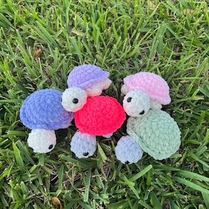 May include: Five crocheted turtle toys in different colors: lavender, light blue, pink, hot pink, and light green. The turtles are sitting on a bed of green grass.
