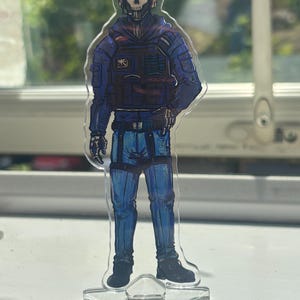 Puede incluir: Una figura de acrílico transparente con la ilustración de un personaje que lleva una máscara de calavera, una sudadera táctica azul, un chaleco oscuro y pantalones azules. El personaje tiene auriculares y se apoya en una base de estrella.