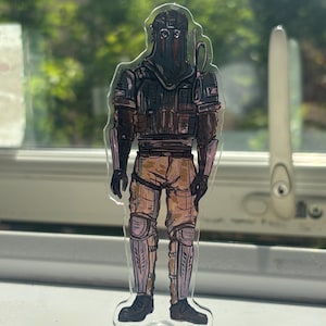 May include: A clear acrylic standee featuring a character illustration of a figure wearing a dark tactical helmet, a dark vest, and tan trousers with knee pads. The figure stands on a transparent star-shaped base, showcasing detailed line art.