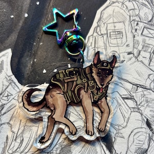 May include: A clear acrylic charm featuring a cartoon dog in a military-style vest and cap. The dog is brown and tan with a red tongue. The charm has a star-shaped iridescent clasp and a metal ring.