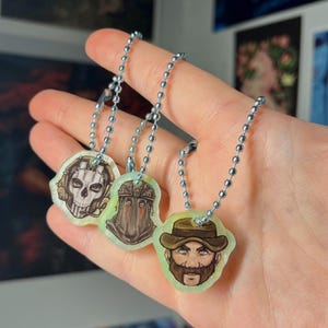 May include: Three character pendants with ball chain necklaces. The pendants feature detailed illustrations of a skull with headphones, a masked figure, and a bearded man wearing a hat. The pendants are on a light green background.
