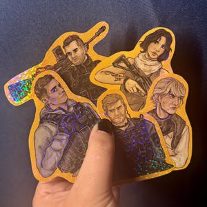 May include: A collection of five holographic stickers featuring illustrations of characters with weapons. The stickers have a gold border and are held in a hand against a blue background. The characters are depicted in tactical gear.