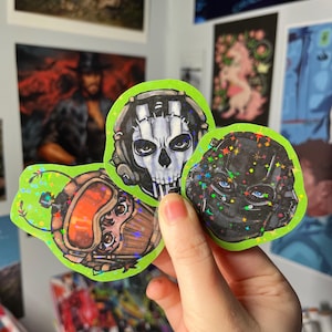 Könnte beinhalten: Drei holografische Aufkleber mit Charakterdesigns. Einer zeigt einen Schädel mit Headset, ein anderer einen schwarzen Helm mit blauen Augen und der dritte eine orange getönte Maske. Jeder Aufkleber hat einen grünen Rand.