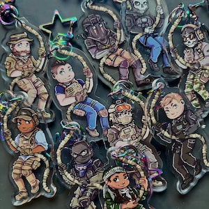 May include: A selection of clear acrylic keyrings showcasing cartoon illustrations of soldiers in combat attire. Each keyring features a unique design, with characters holding a rope. The keyrings have iridescent hardware.