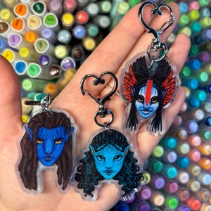 Puede incluir: Tres llaveros temáticos de Avatar con personajes de piel azul con rasgos faciales y peinados detallados. Cada llavero tiene un cierre plateado en forma de corazón. El fondo está lleno de marcadores de colores.