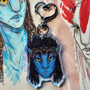 Puede incluir: Un llavero acrílico transparente con un personaje de piel azul con ojos amarillos y trenzas negras. El llavero tiene un cierre plateado en forma de corazón. El personaje es de la película Avatar. El fondo muestra otros bocetos de personajes de Avatar.