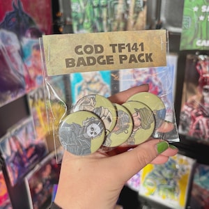 Puede incluir: Una bolsa de plástico transparente que contiene un paquete de insignias COD TF141. El paquete incluye varias insignias redondas de color verde oliva con ilustraciones de personajes. El texto "COD TF141 BADGE PACK" está impreso en el embalaje. Las insignias se sostienen en una mano.