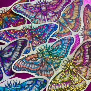 May include: A collection of colorful moth stickers with iridescent glitter accents. The stickers feature detailed illustrations in shades of purple, blue, orange, and yellow. The stickers are arranged on a purple glitter surface.