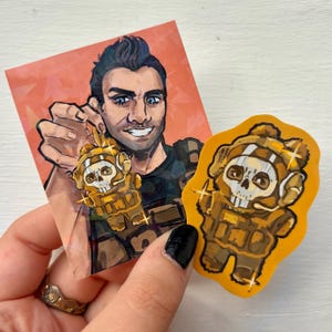 May include: An illustration of a man holding a gold-colored charm with a skull design. Next to it is a sticker of the same charm. The charm and sticker have a military theme, with a skull wearing a helmet and tactical gear.