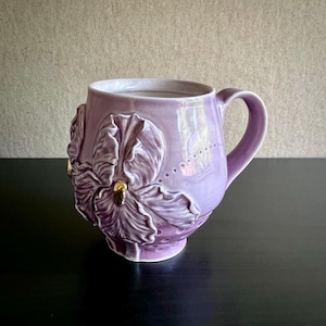 May include: A lilac ceramic mug with an embossed floral design. The mug has a curved handle and gold accents on the flower's center. The floral design is repeated on the mug's surface, giving it a textured appearance. The mug is suitable for hot beverages.