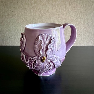 May include: A lilac ceramic mug featuring a raised floral design with detailed petals and a gold-coloured centre. The mug has a curved handle and a smooth, glossy finish. The mug is on a black surface.