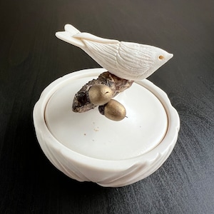 May include: A white ceramic trinket box with a carved bird perched on a branch with two golden acorns. The bird is detailed with carved feathers and a small eye. The box has a lid and a textured, scalloped base.