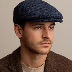 May include: A navy blue herringbone pattern flat cap. The cap is made of a textured fabric and has a rounded crown and a small, stiff brim. The cap is worn by a person wearing a brown blazer.