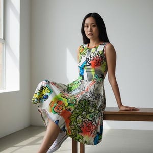 Op de afbeelding: Een mouwloze, knielange jurk met een levendige graffiti-achtige print in groen, oranje, blauw en geel. De jurk wordt gedragen met witte sneakers. Het model zit op een houten bank in een lichte kamer.