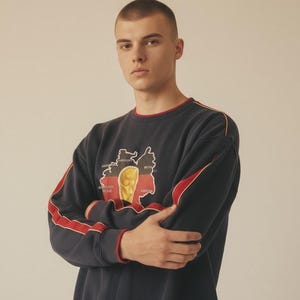 May include: Navy blue sweatshirt with red and yellow accents. The sweatshirt features a graphic of a map with a gold trophy. Red stripes run down the sleeves. The collar and cuffs are trimmed in red.