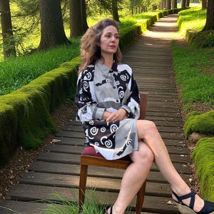 May include: A woman wearing a black, white, and gray patterned dress and black sandals sits on a wooden chair. The dress features a swirling design. The background shows a wooden walkway surrounded by green grass and trees.