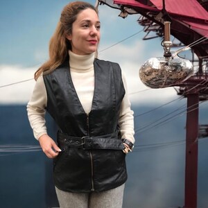 May include: A woman wearing a black leather vest with a zipper closure and a belt. The vest has a v-neck and is worn over a white turtleneck sweater. The woman is standing in front of a blue and white background.