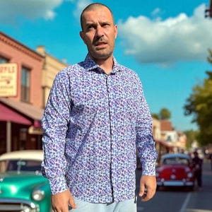 May include: A button-down shirt with a floral pattern in shades of purple and blue. The long-sleeved shirt is worn with light blue pants. The background features a street scene with classic cars and buildings under a bright blue sky.