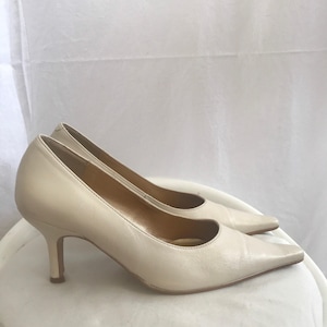 May include: A pair of cream-coloured leather court shoes with pointed toes and a mid-height heel. The shoes are set on a white surface, showcasing their elegant design. The heels measure approximately 6.35 cm.