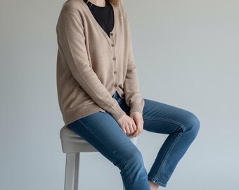 May include: A woman wearing a tan cardigan over a black shirt and blue jeans, sitting on a white stool. The cardigan has buttons down the front. White sneakers complete the outfit. The background is a solid light gray.