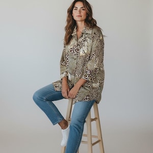 Può includere: Una donna indossa una camicia abbottonata con una stampa animalier mista di leopardo e zebra. Indossa jeans blu e scarpe da ginnastica bianche, seduta su uno sgabello di legno. La camicia ha un colletto e maniche a tre quarti.