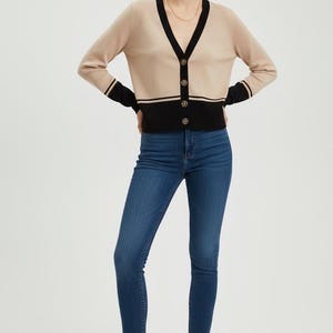 Puede incluir: Un cárdigan beige con ribete negro y botones dorados, combinado con vaqueros pitillo azul oscuro. El cárdigan tiene cuello en V y mangas largas con rayas negras en los puños. Los vaqueros son de cintura alta y ajustados.