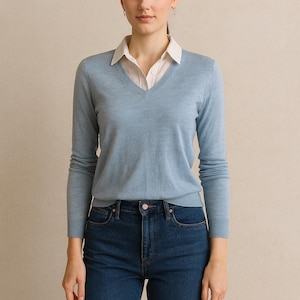 Könnte beinhalten: Hellblauer V-Ausschnitt-Pullover über einem weißen Hemd mit Kragen, kombiniert mit dunkelblauen Jeans. Das Outfit ist schlicht und klassisch, geeignet für Freizeit- oder Business-Casual-Anlässe. Das Model steht vor einem neutralen Hintergrund.