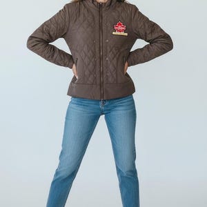 Może przedstawiać: Brązowa pikowana kurtka z zapięciem na zamek i małą czerwoną i złotą odznaką na piersi. Kurtka jest połączona z niebieskimi dżinsami i białymi trampkami. Kurtka ma wzór w romby.