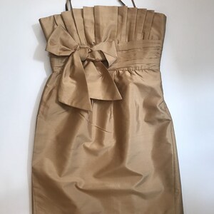 Puede incluir: Un vestido dorado sin mangas con un gran lazo en la cintura. El vestido tiene un corpiño plisado y una falda ajustada.