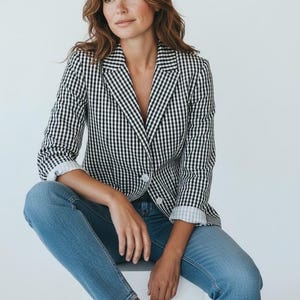 May include: A black and white gingham blazer with a single-button closure. The blazer is paired with blue jeans and white sneakers. The sleeves are rolled up, revealing a white cuff. The outfit is casual and stylish.