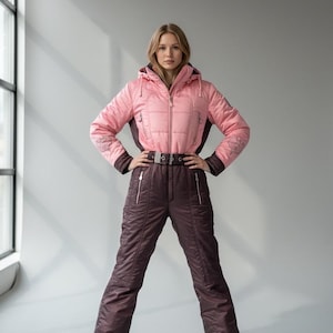 Puede incluir: Un mono de esquí rosa y marrón con una chaqueta con capucha y pantalones a juego. La chaqueta tiene cierre de cremallera y cinturón en la cintura. Los pantalones tienen bolsillos con cremallera. El mono se usa con zapatillas negras.