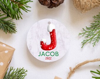 May include: Round white ornament with a red candy cane letter J with white frosting on top. The ornament has a marbleized background and the name "JACOB" in green text and the year "2022" in red text.