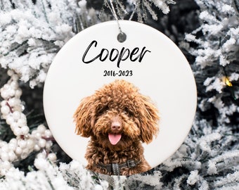 May include: A white ceramic Christmas ornament featuring a brown dog with the name "Cooper" and the years "2016-2023" in black script. The dog has a fluffy coat and is wearing a black collar. The ornament is hanging on a snowy Christmas tree.