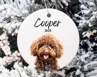 May include: White ceramic Christmas ornament with a brown poodle dog with a pink tongue sticking out. The ornament has the name "Cooper" and the year "2023" written on it.