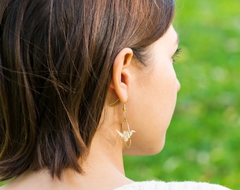 May include: Gold-colored dangle earrings featuring a white origami crane design. The earrings have a delicate, openwork design with a small, circular accent. The background is a blurred green, suggesting an outdoor setting. The earrings are worn on a person.