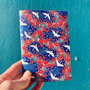 May include: A small notebook with a blue cover featuring a repeating pattern of white cranes flying over pink and blue flowers. The background is a dark blue with a wave pattern.