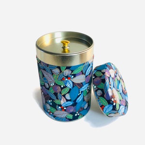 Op de afbeelding: Een ronde metalen theedoos met een gouden deksel en een bloemenpatroon. De doos is versierd met blauwe, groene, paarse en gouden bladeren en bessen. Het deksel heeft een gouden knop.