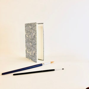 May include: A white book with a black and white wave pattern on the cover. The book is closed and standing upright on a white surface. Two blue paintbrushes are lying on the surface in front of the book.