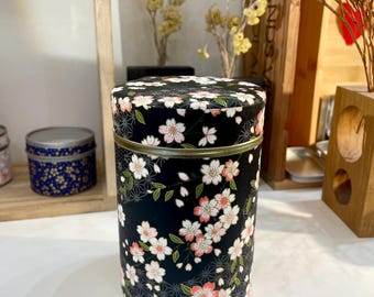 May include: A black cylindrical tin with a floral pattern of pink cherry blossoms and green leaves on a black background. The tin has a gold rim.