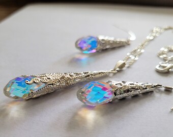 May include: A silver necklace and earrings set with iridescent teardrop-shaped crystals. The crystals have a rainbow effect and are set in ornate silver filigree.