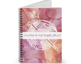Pode incluir: Um caderno de espiral com um design de mármore rosa e dourado. A capa apresenta um contorno branco de uma bola de fio e um gancho de crochê com o texto "Crochet is my happy place!"