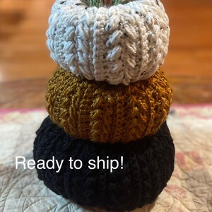 May include: A stack of three crocheted pumpkins in cream, gold, and black. The top pumpkin has a green stem and leaves. The pumpkins are arranged vertically, creating a fall-themed decorative item.