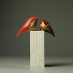 Op de afbeelding: Twee houten vogelsculpturen, een rood en goud, de andere bruin en goud, op een wit houten voetstuk.