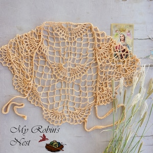 May include: A light peach-colored, open-weave crochet shawl with a delicate, lacy pattern. The shawl has a rounded shape and tie closures. A small religious image and decorative grasses are in the background. The text "My Robin's Nest" is at the bottom.