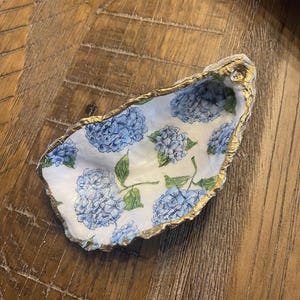 May include: A white oyster shell with a gold rim, decorated with a blue and green floral pattern. The shell is a decorative dish or trinket holder.