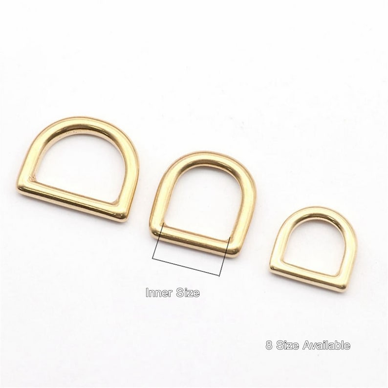 Op de afbeelding: Drie gouden metalen D-ringen in verschillende maten. De ringen zijn gerangschikt van groot naar klein. De tekst "Inner Size" staat onder de middelste ring. De tekst "8 Size Available" staat onder de kleinste ring.