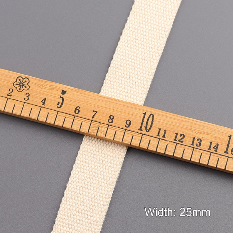 May include: A close-up of a natural-colored cotton webbing measuring 25mm wide. The webbing is laid on top of a wooden ruler with markings in centimeters.